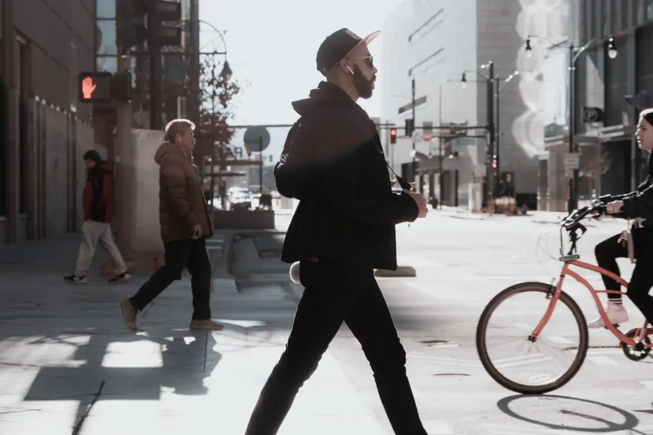 A man in a cap walks across a sunlit street, embodying the best focal length for street photography. People on bikes and sidewalks fill the urban scene.