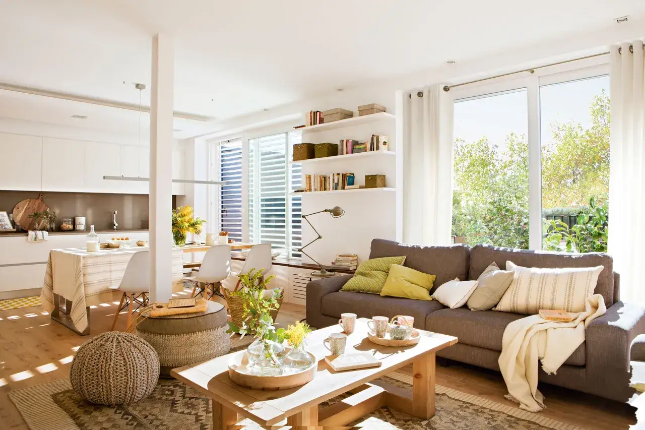 Espaciosa distribuci&oacute;n loft con cocina abierta, comedor y sala de estar. Sof&aacute; gris, mesa de centro de madera, alfombra y mucha luz natural.