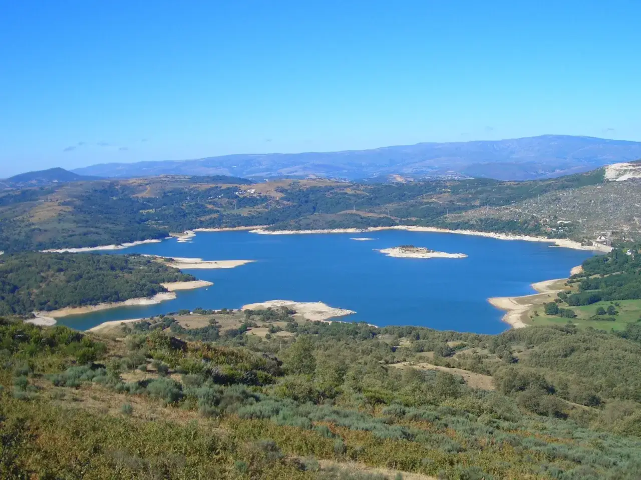 El embalse de Prada, con su agua azul brillante, se extiende entre colinas verdes y secas bajo un cielo despejado.