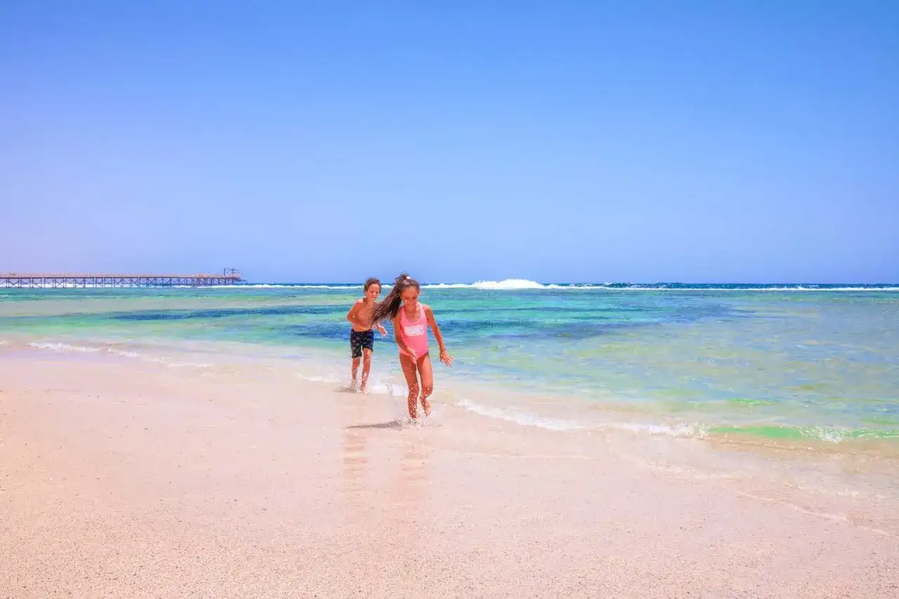 Dzieci biegają po plaży w Marsa Alam, woda jest krystalicznie czysta, a w oddali widać pomost. Idealne wakacje z pickalbatros.
