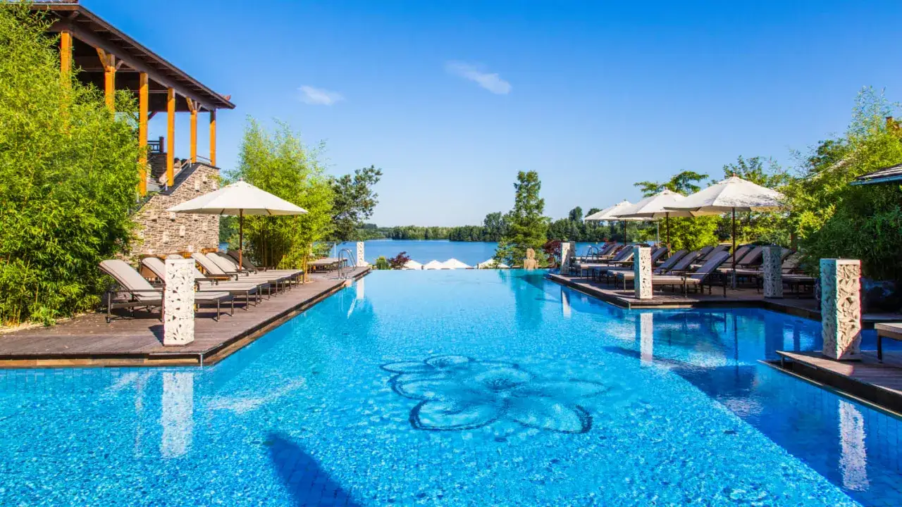 Ein luxuriöser Pool mit Blick auf einen See lädt zum Schwimmen in NRW ein. Sonnenliegen und Bambus säumen das blaue Wasser.