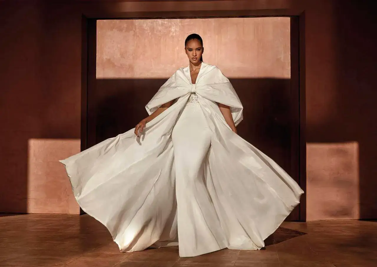Une femme porte une magnifique tenue de mariage religieux, une robe blanche évasée avec une cape spectaculaire.