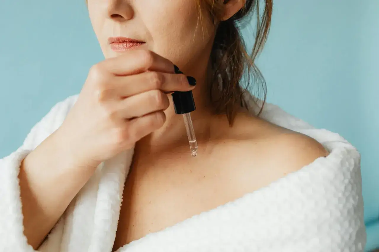 Kobieta w szlafroku aplikuje serum z pipetki na dekolt. Zastanawiasz się retinol co to? To składnik odmładzający skórę.
