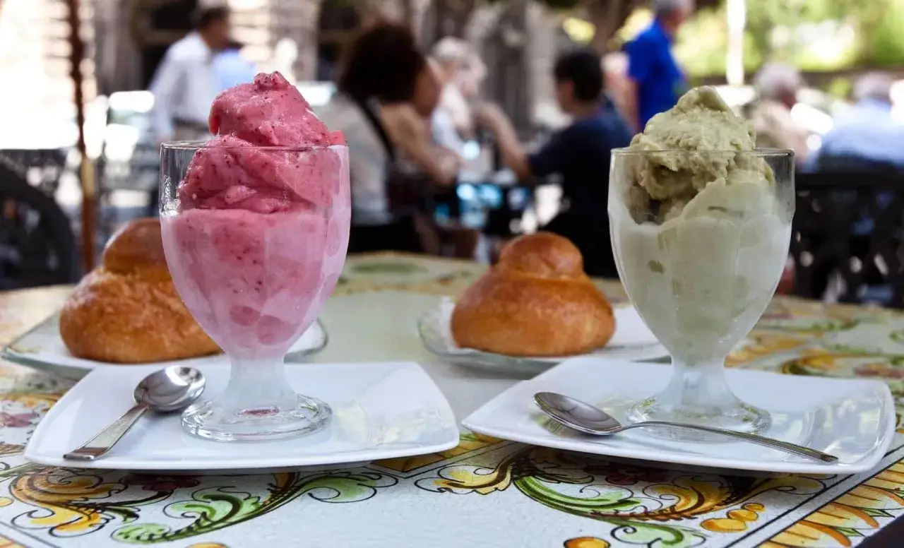 Tv&aring; glas med glass, en rosa och en gr&ouml;n, st&aring;r p&aring; ett bord med m&ouml;nstrad duk. I bakgrunden syns m&auml;nniskor p&aring; en uteservering, kanske p&aring; en av alla mysiga restauranger i Aten.