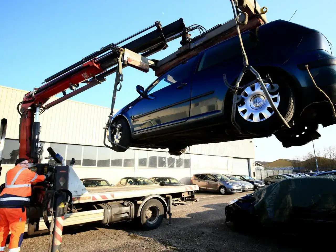 Un ouvrier en combinaison orange utilise une grue pour soulever une voiture bleue, probablement suite à une amende pour stationnement gênant.