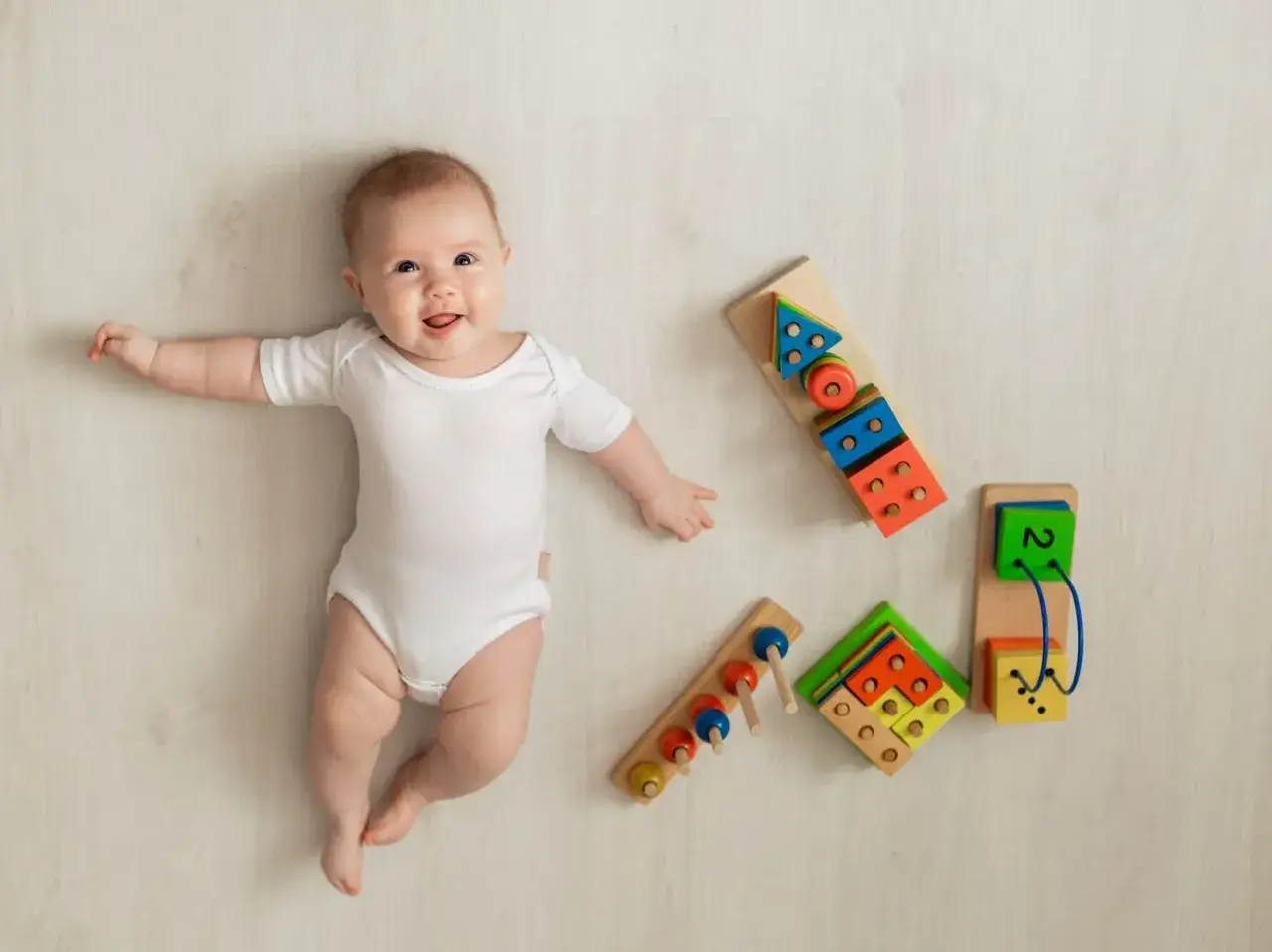 Uśmiechnięte 5 miesięczne dziecko w białym body leży obok drewnianych zabawek edukacyjnych.