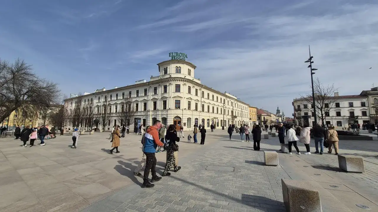 Ludzie spacerują po jasnym, przestronnym placu. W tle widać hotel Europa i zabytkowe kamienice.