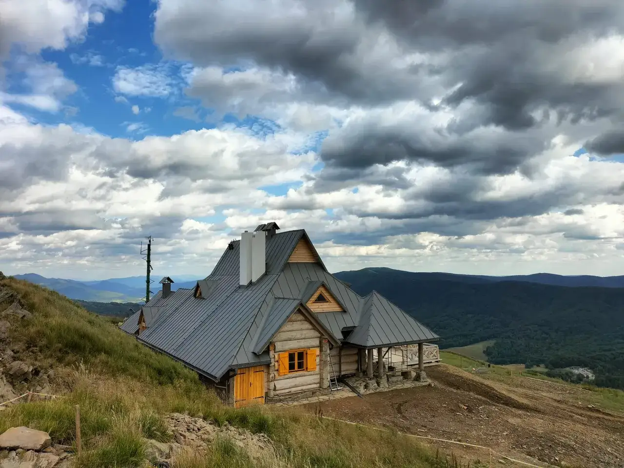Drewniany dom na zboczu g&oacute;r, z widokiem na malownicze Bieszczady. Chmury na niebie dodają dramatyzmu krajobrazowi.