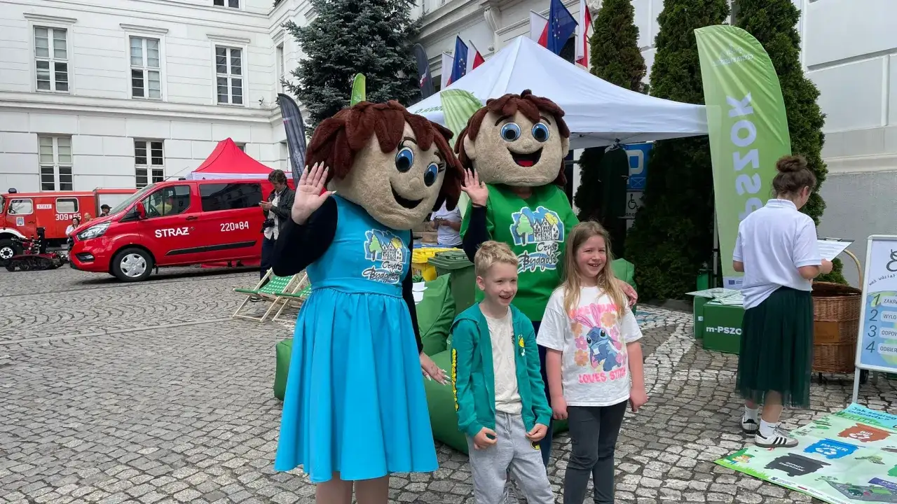 Dwa pluszowe misie i dzieci pozdrawiają na Dzień Dziecka. W tle czerwony wóz strażacki i namioty.