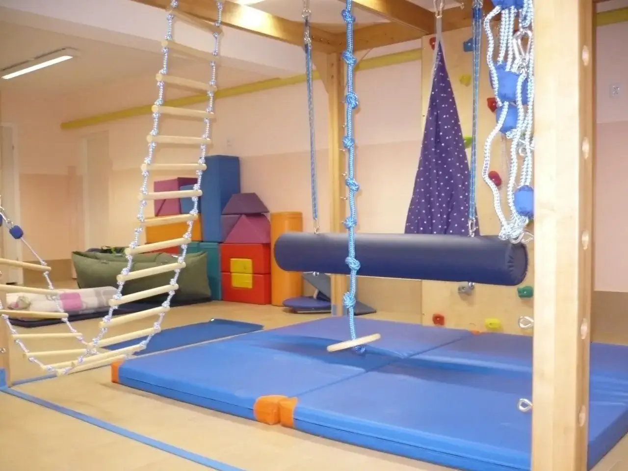 Kolorowa sala integracji sensorycznej z drabinką, huśtawkami i miękkimi materacami.
