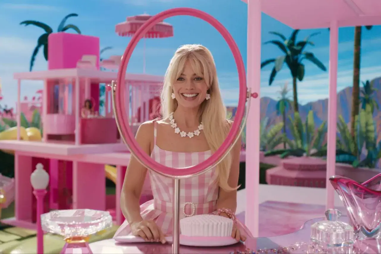 Margot Robbie als Barbie, lächelnd durch einen pinken Spiegel, in einer rosa karierten Kleidung, vor einer pinken Villa.
