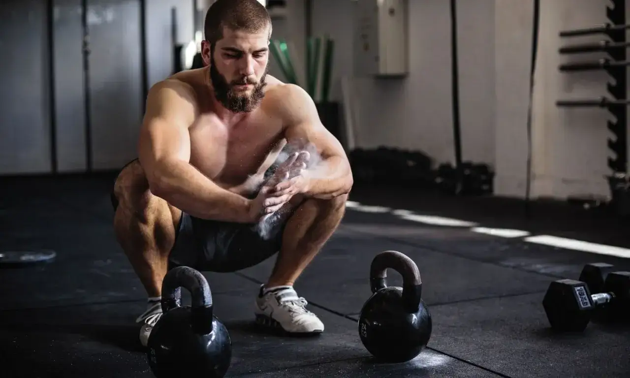 Uomo con barba si prepara per un allenamento funzionale palestra cos'è, con kettlebell e manubri sullo sfondo.