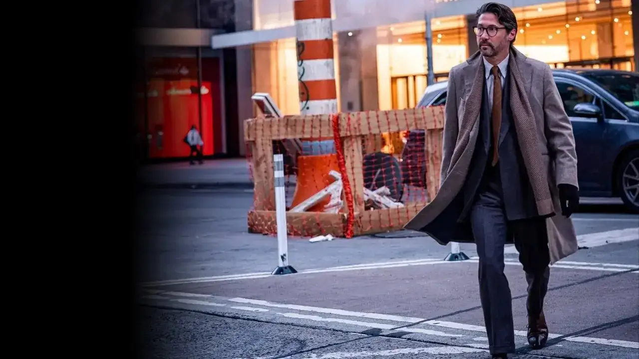Uomo con elegante stile outfit, cappotto lungo, completo grigio e cravatta marrone cammina per strada.