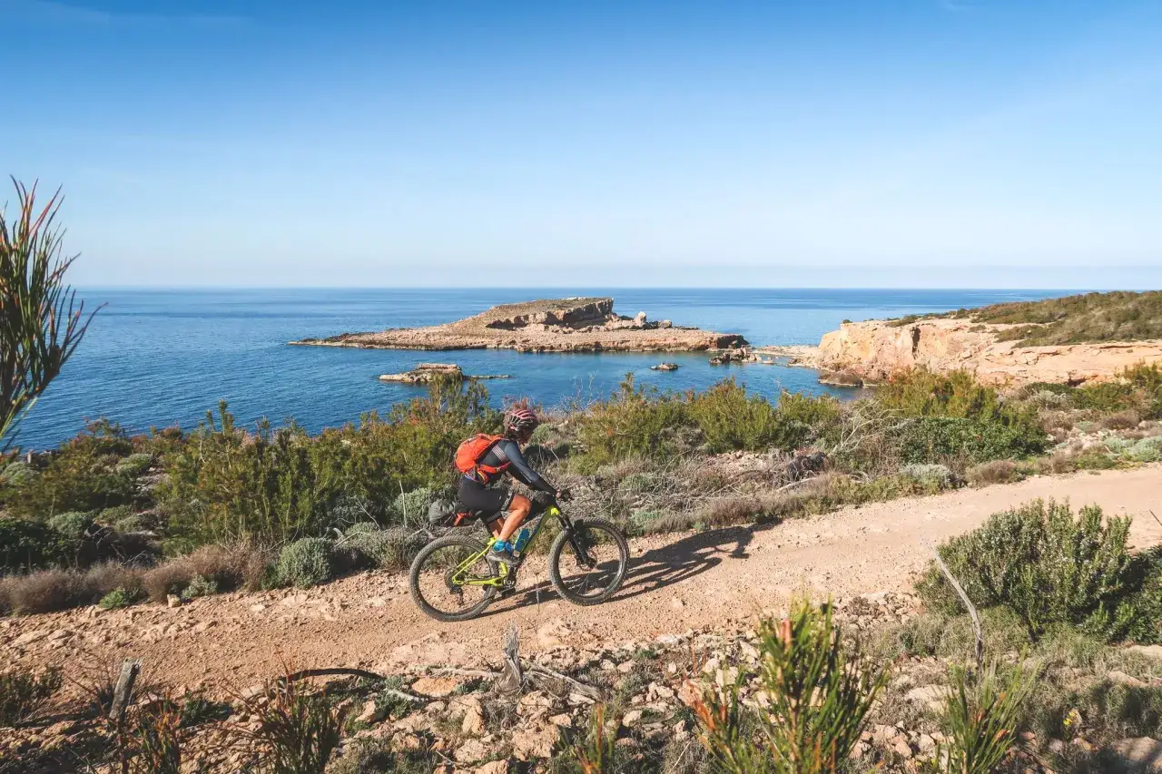 Ciclista disfruta de un recorrido por la costa de Ibiza en bicicleta, con el mar azul y rocas al fondo.