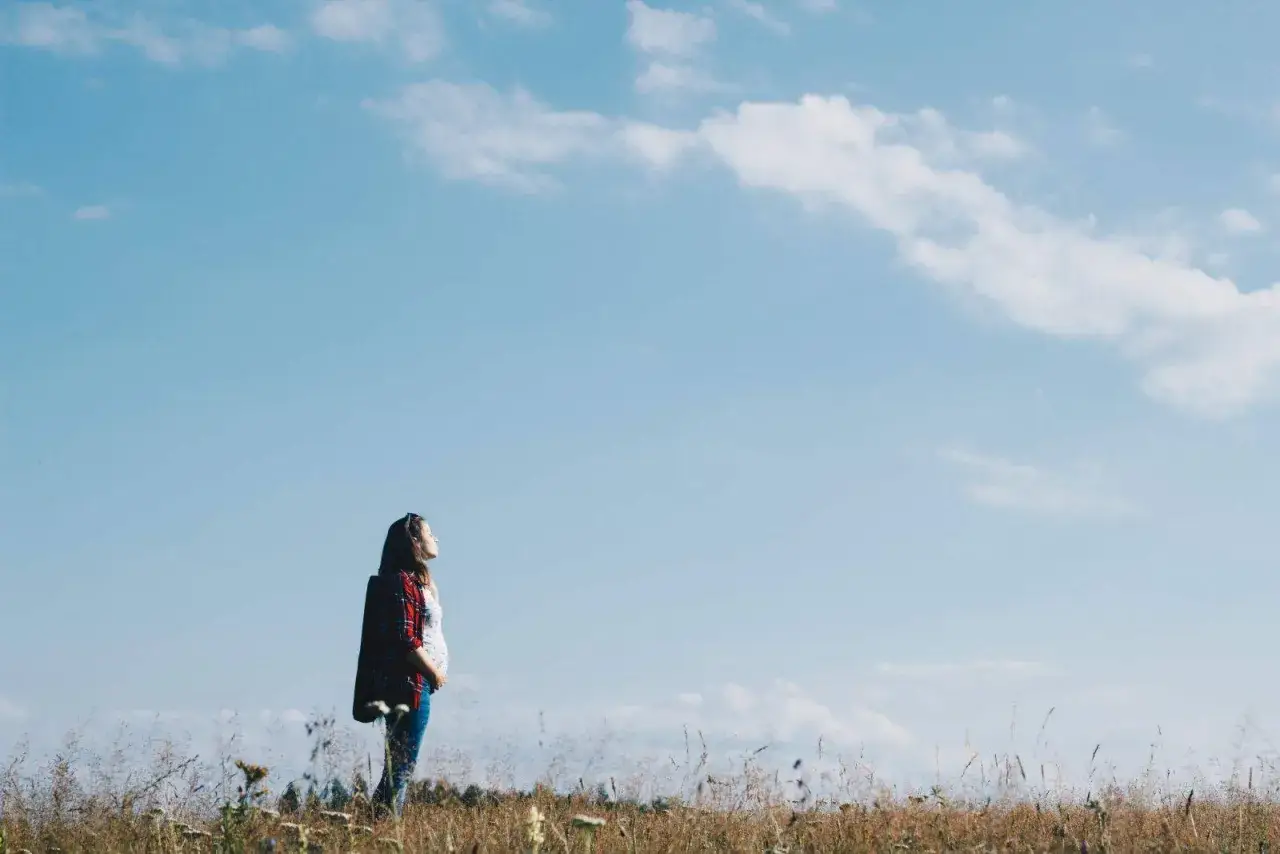 Donna in piedi in un prato, con la pancia visibile, sotto un cielo azzurro. Sembra una montagna a 1500 metri, con un senso di pace e attesa.