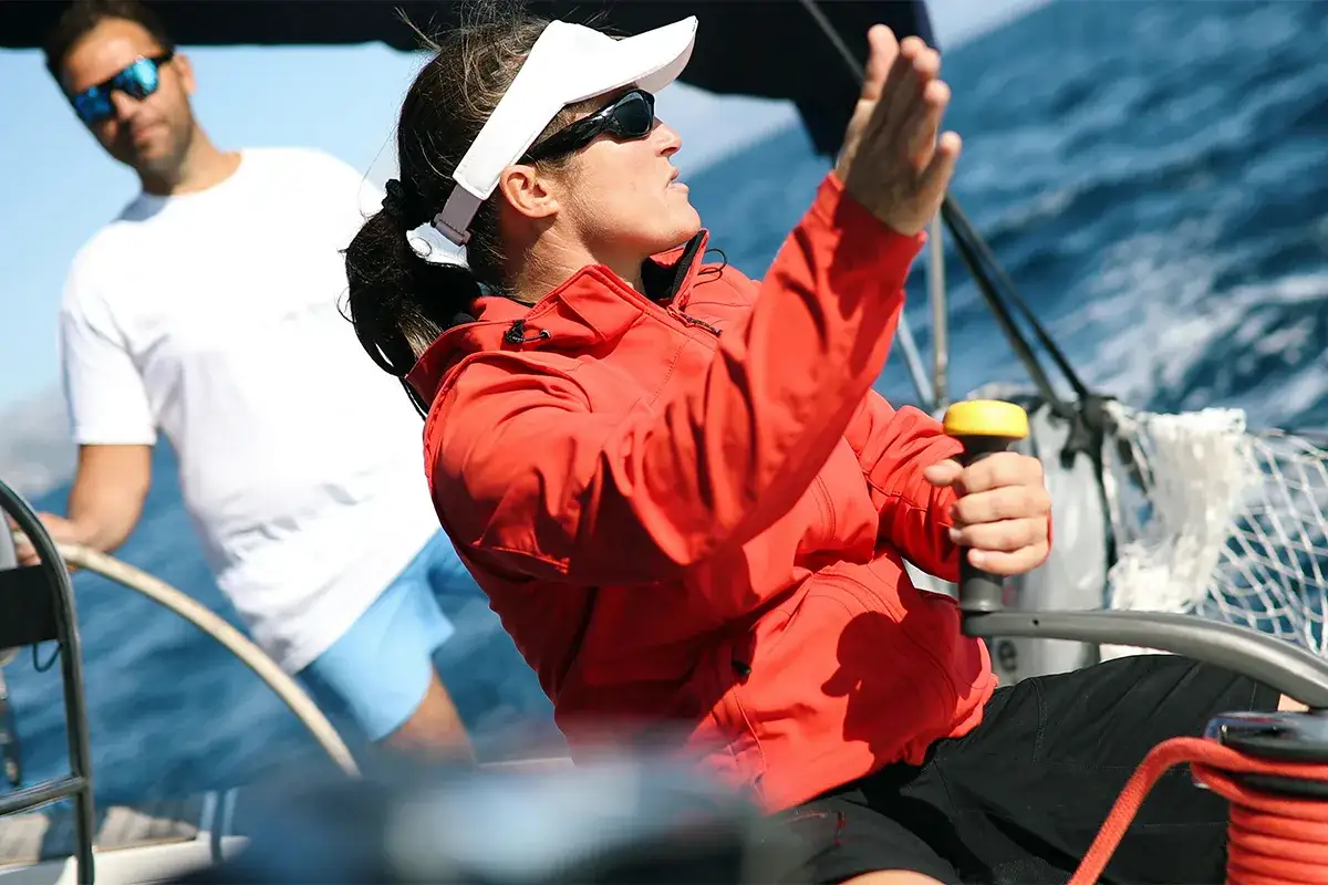 Une femme en veste rouge, le gouvernail en main, dirige le voilier. Elle est la personne qui conduit un bateau. Un homme en arri&egrave;re-plan.
