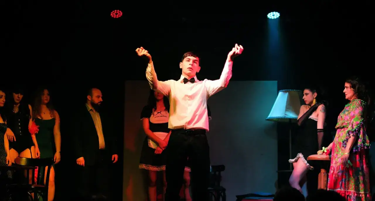Un giovane attore, con le braccia alzate, sembra essere il bersaglio di uno sguardo intenso. Attorno a lui, altre figure femminili e un uomo completano la scena teatrale.