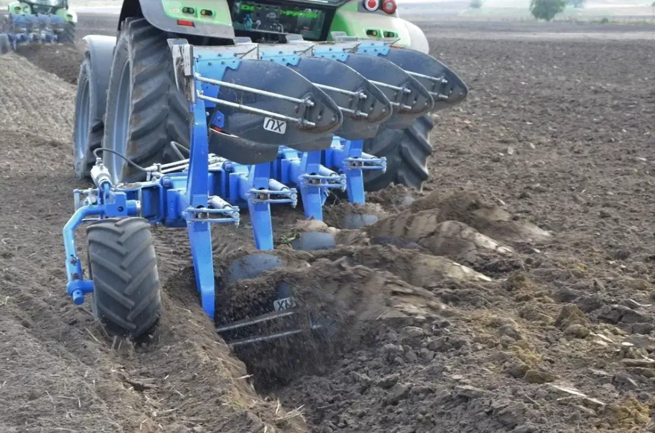 Traktor z pługiem orze pole, odwracając ziemię. Widać pracujące lemiesze i koło podporowe.