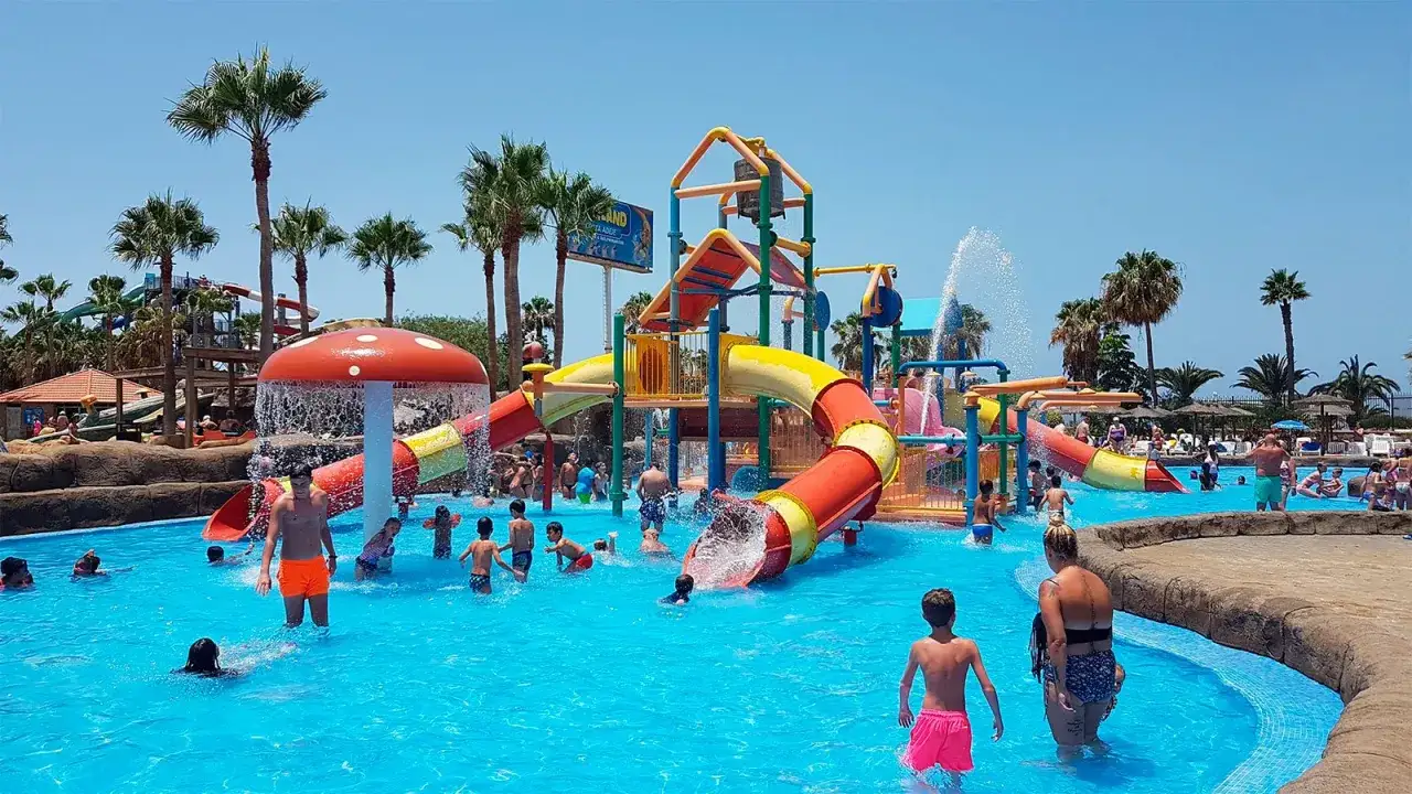 Niños y adultos disfrutan de toboganes y piscinas en un día soleado en el mejor parque acuático de Europa. Palmeras y juegos acuáticos crean un ambiente festivo.