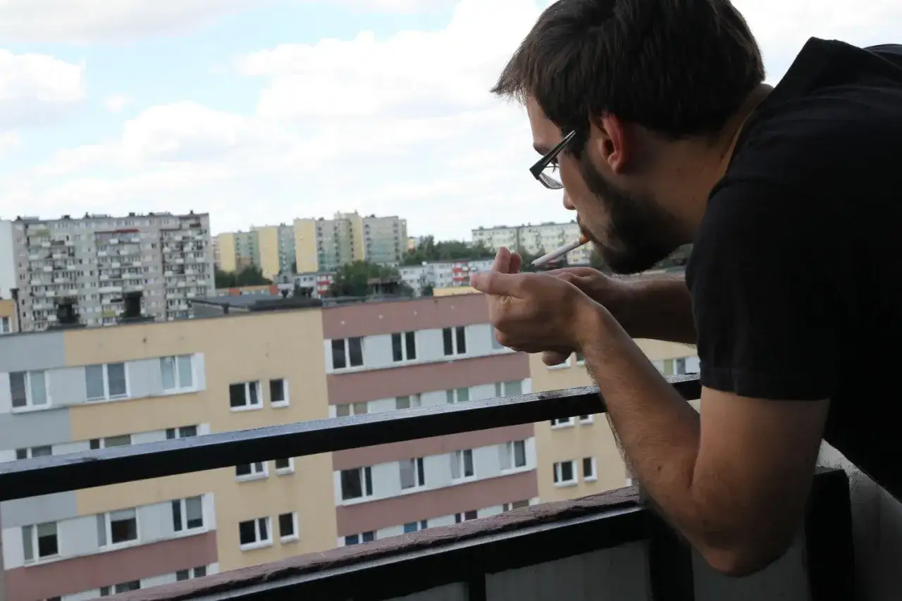 Mężczyzna z brodą i okularami zapala papierosa na balkonie. W tle widać blokowisko. Sąsiad pali na balkonie.