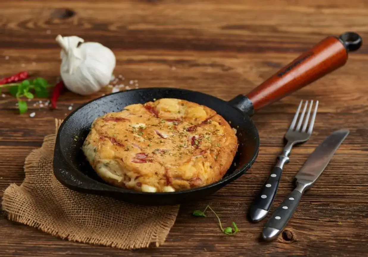 Słony omlet z szynką i serem na patelni, idealny przepis na szybkie śniadanie. Obok widelec i nóż.