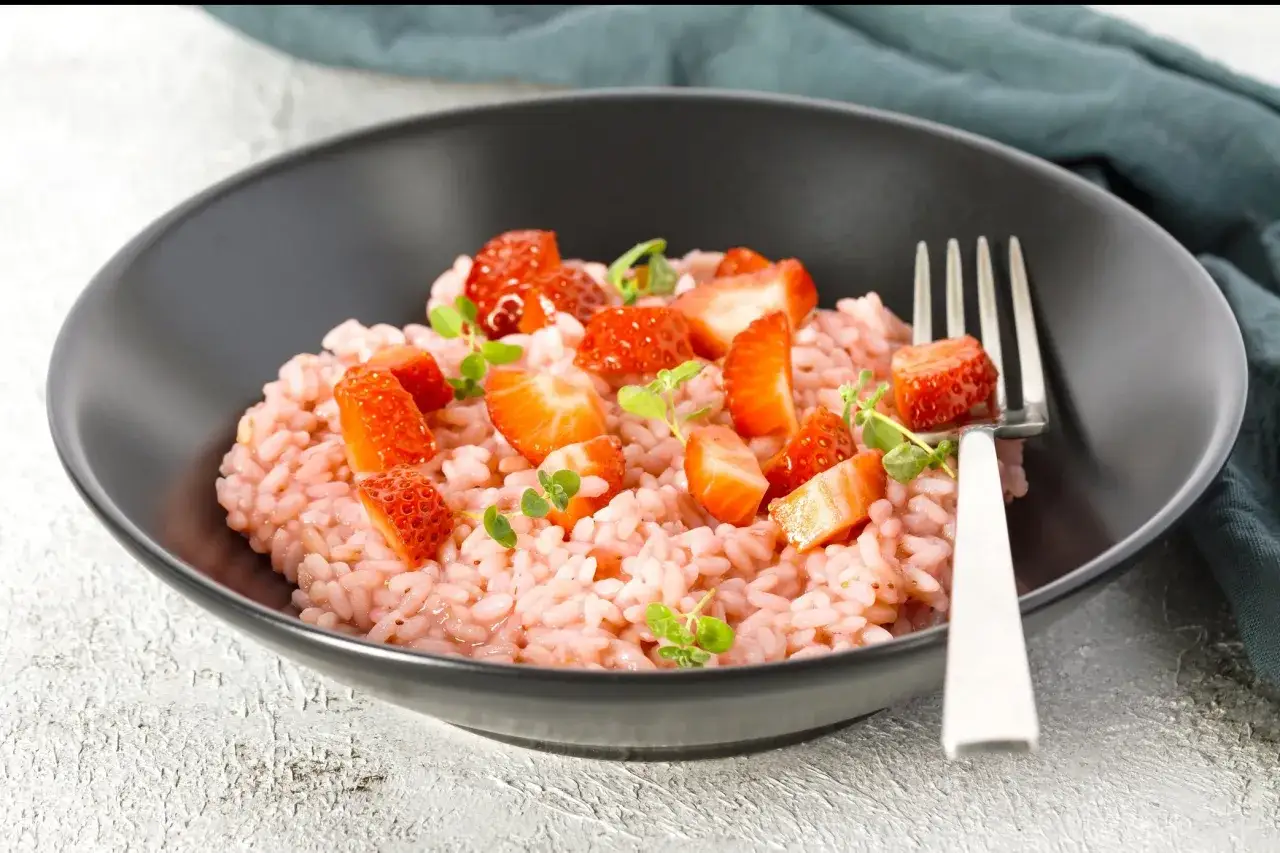 Un cremoso risotto alle fragole, guarnito con fragole fresche a pezzi e germogli verdi, servito in una ciotola scura con una forchetta.