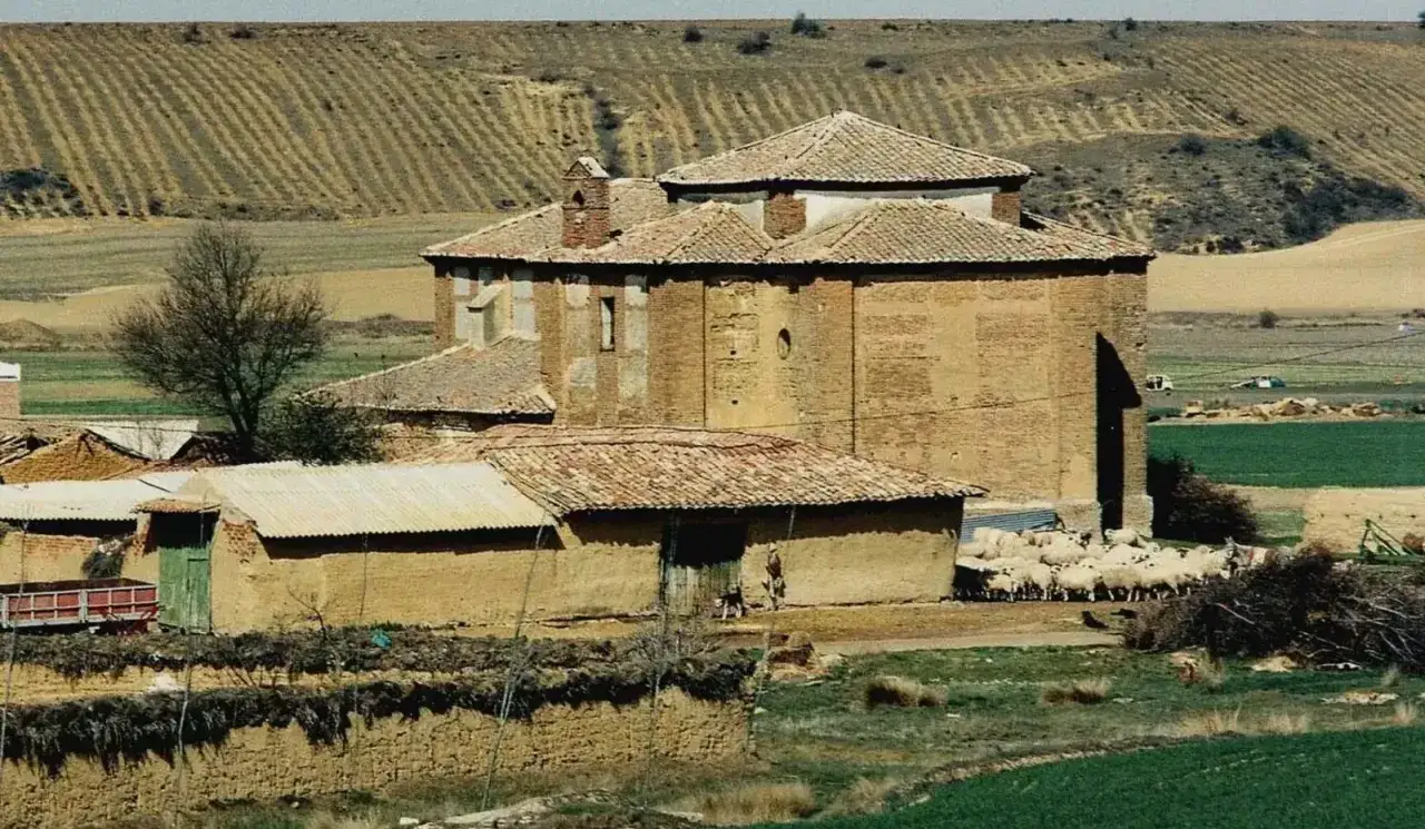 Edificio de ladrillo en Calzadilla de la Cueza, con rebaño de ovejas pastando cerca. Campos labrados al fondo.