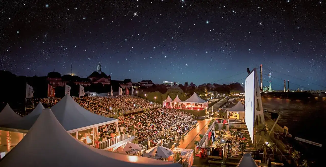 Ein belebtes Open-Air-Kino im Ruhrgebiet unter einem sternenklaren Himmel. Tausende Besucher sitzen auf Tribünen, umgeben von Zelten und der Kulisse einer Stadt am Fluss.