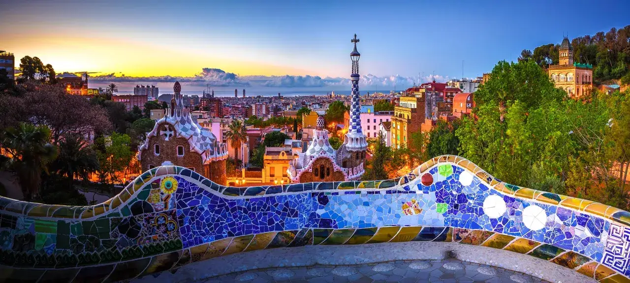 Bunte Mosaikb&auml;nke im Vordergrund, dahinter Gaud&iacute;s M&auml;rchenh&auml;user und die Skyline von Barcelona bei Sonnenuntergang. Ein atemberaubender Blick auf Barcelona von oben.