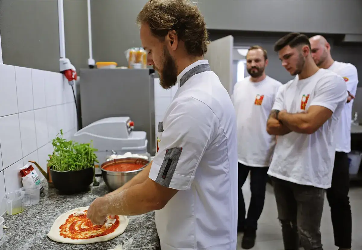 Pizzaiolo, mistrz pizzy, przygotowuje ciasto. W tle stoją jego koledzy, obserwując proces.