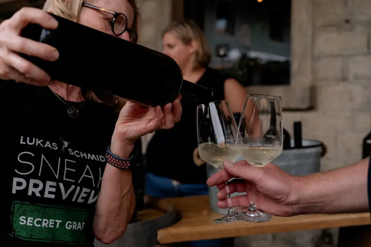 Eine Frau schenkt Scheurebe Wein in Gläser ein. Ein Mann hält zwei Gläser mit dem hellgelben Wein.