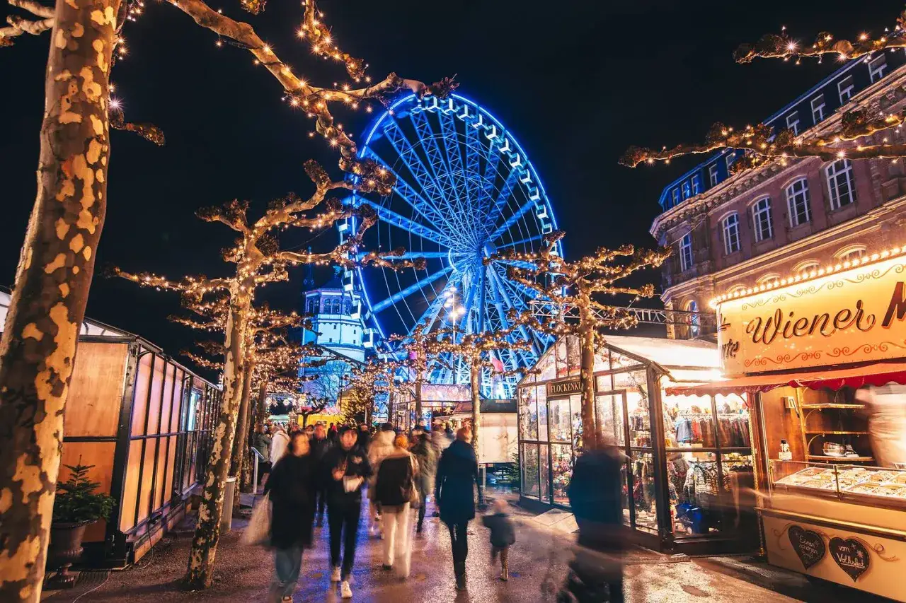Menschen schlendern über einen stimmungsvollen Weihnachtsmarkt in NRW, beleuchtet von Lichterketten und einem Riesenrad. Ein echter Geheimtipp!