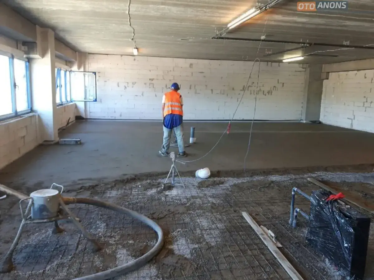 Robotnik w kamizelce nadzoruje prace przy wylewce z miksokreta. Na podłodze widać świeży beton, siatkę zbrojeniową i sprzęt budowlany.