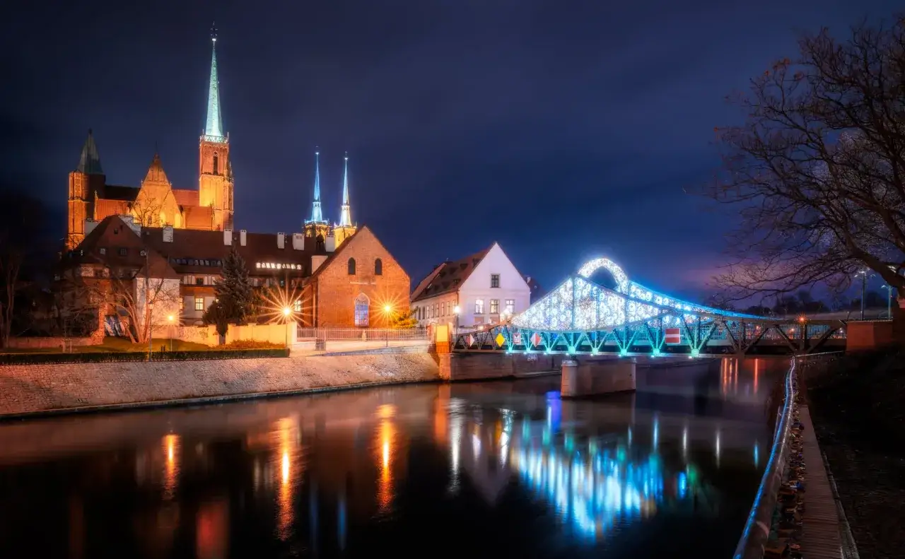 Nocny Wrocław zachwyca iluminacjami. Katedra i most Tumski, idealne wrocławskie atrakcje dla dorosłych, odbijają się w wodzie.