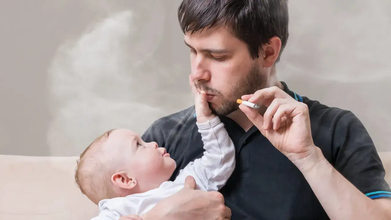 Mann h&auml;lt Baby und raucht in der Wohnung. Rauch umgibt sie. Das Jugendamt k&ouml;nnte eingreifen.