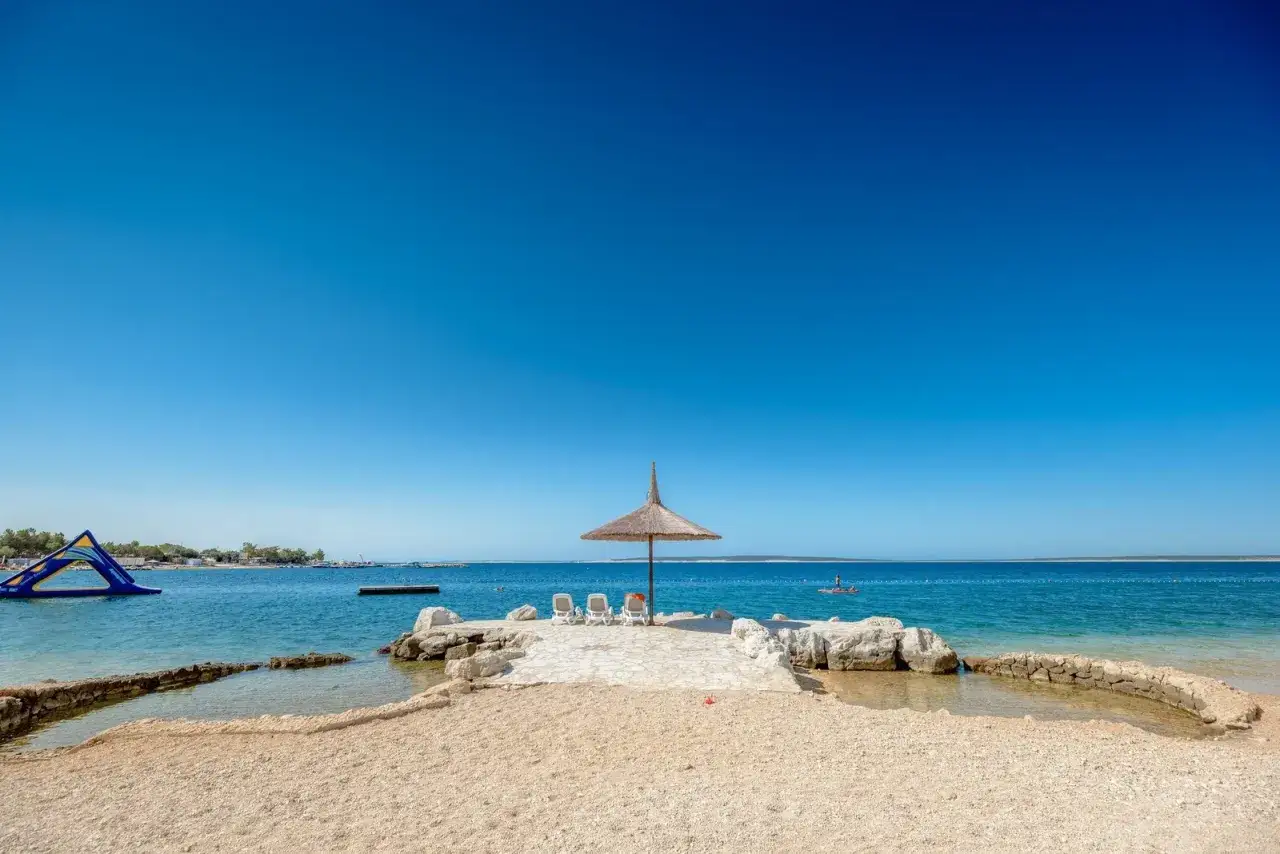 Słoneczna plaża z parasolem i leżakami w camping village &Scaron;imuni. W tle widać wodny plac zabaw i spokojne morze.
