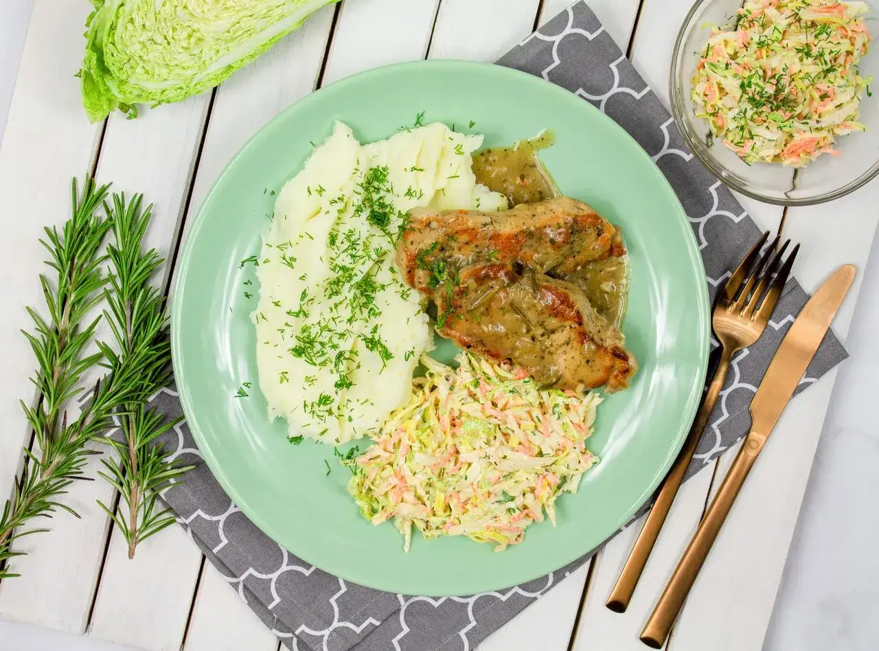 Pyszny schab w sosie podany z puree ziemniaczanym i surówką z kapusty. Całość ozdobiona świeżym koperkiem i gałązkami rozmarynu.