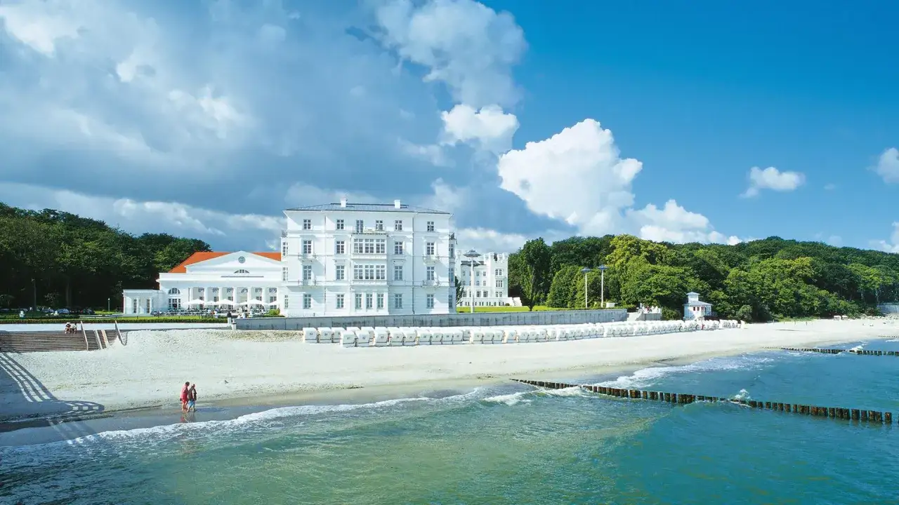 Ein weißes Gebäude mit roten Ziegeldach am heiligendamm strand. Viele Strandkörbe reihen sich am Sand.