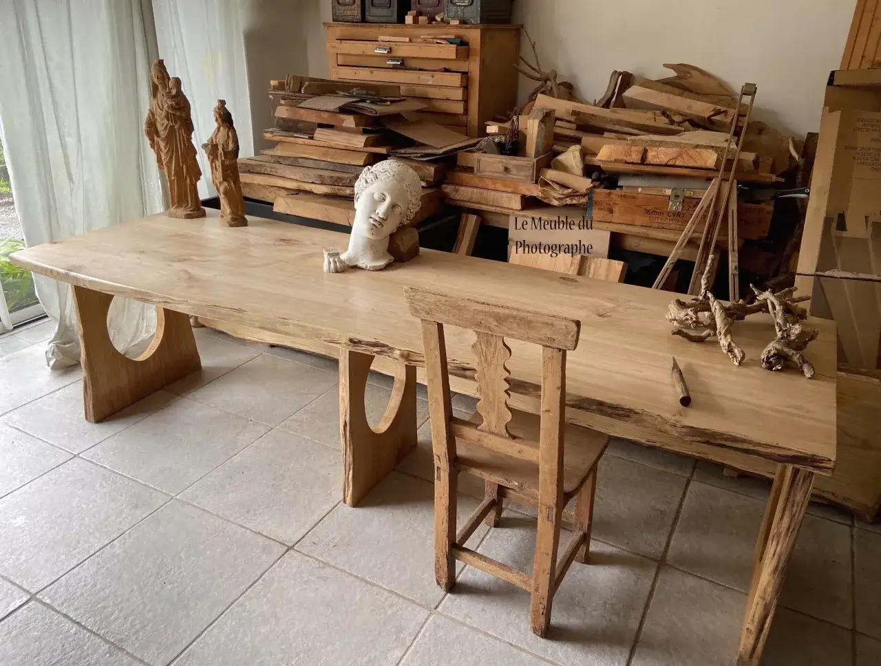 Un atelier de menuiserie avec des planches de bois brut, des sculptures et une table en bois massif. Idéal pour réparer meuble bois.