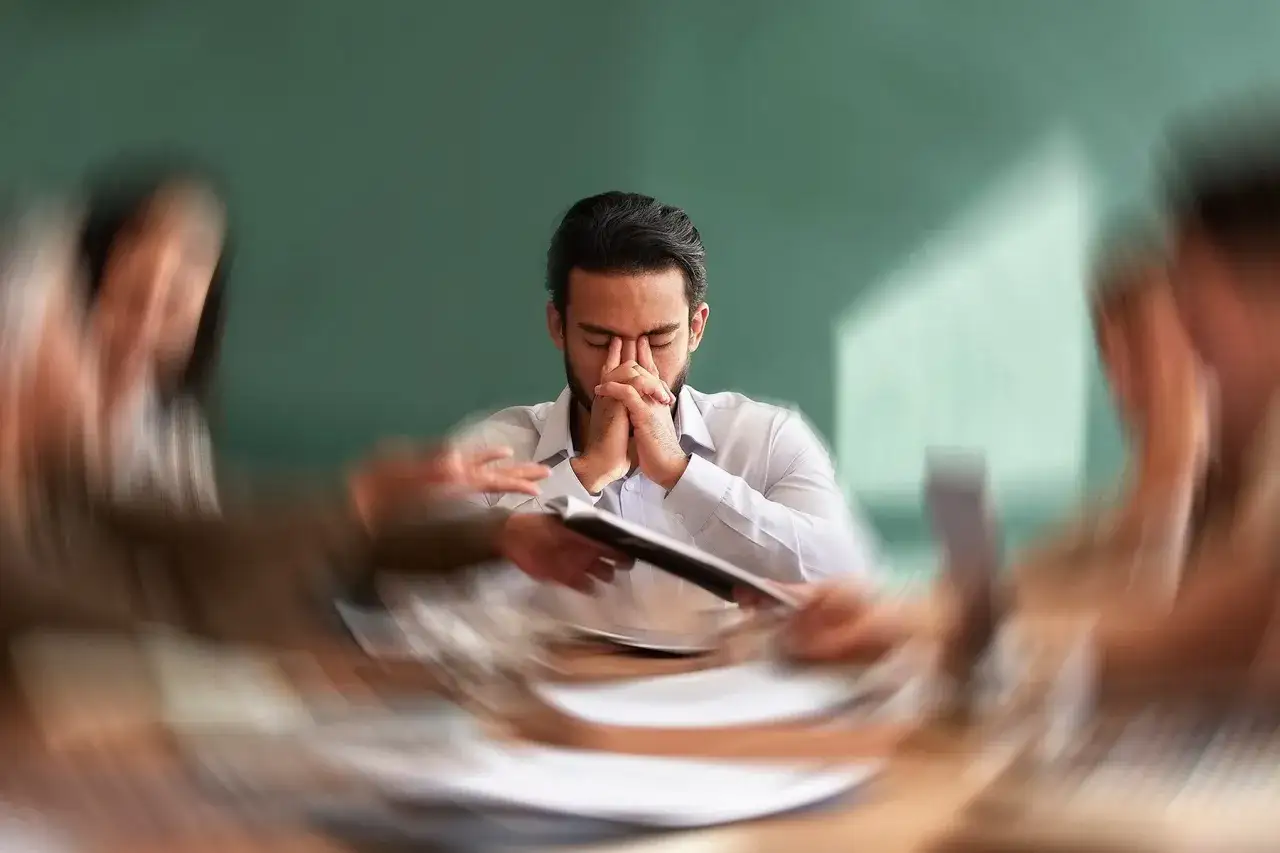 Uomo con le mani sul viso, circondato da figure sfocate, che lotta con l'ansia ad alto funzionamento durante una riunione.
