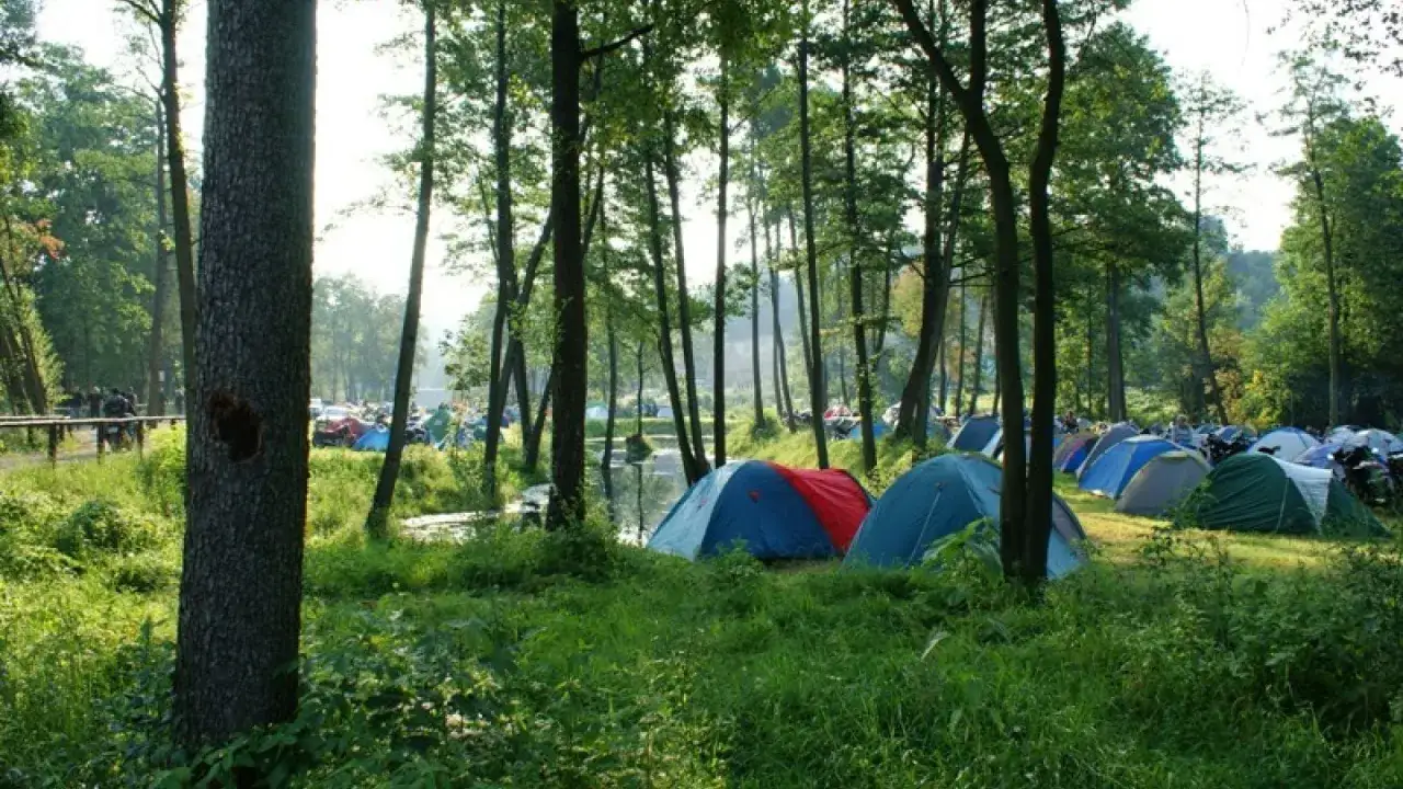 Kolorowe namioty rozstawione w lesie, nad wodą. W tle widać motocykle i ludzi. To idealne miejsce na **camping Świętokrzyskie**.