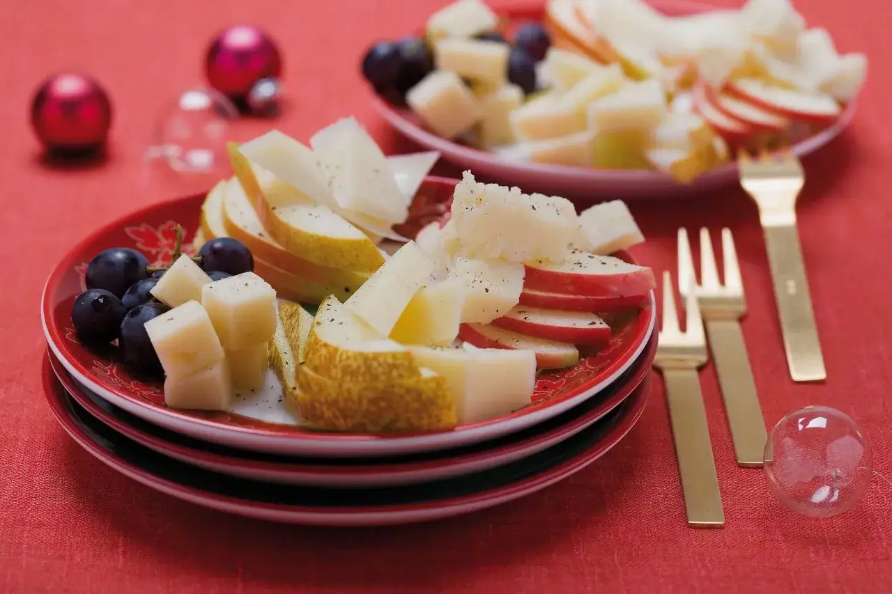 Piatti rossi con formaggi a pasta cotta, pere, mele e uva. Decorazioni natalizie sullo sfondo.