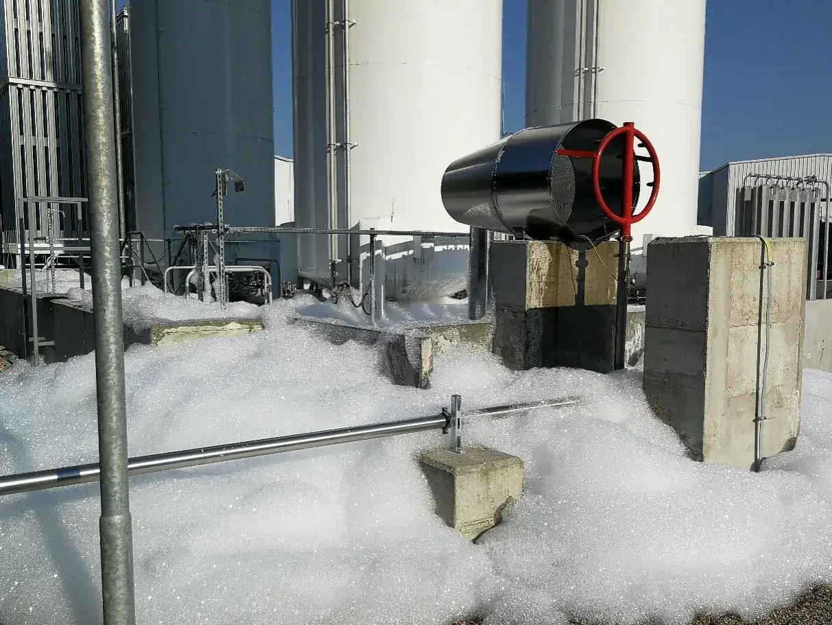 Duże zbiorniki przemysłowe otoczone pianą. Widoczny jest czarny metalowy element z czerwonym uchwytem, który może być częścią systemu kontrolującego liczbę spienienia.