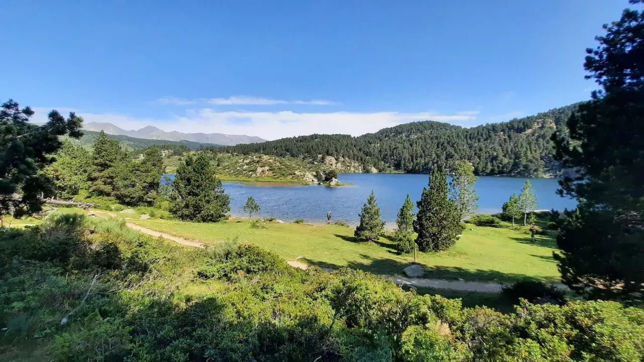 Un lago azul sereno en la **Alta Cerdanya**, rodeado de pinos y monta&ntilde;as. Un excursionista solitario pasea por la orilla.