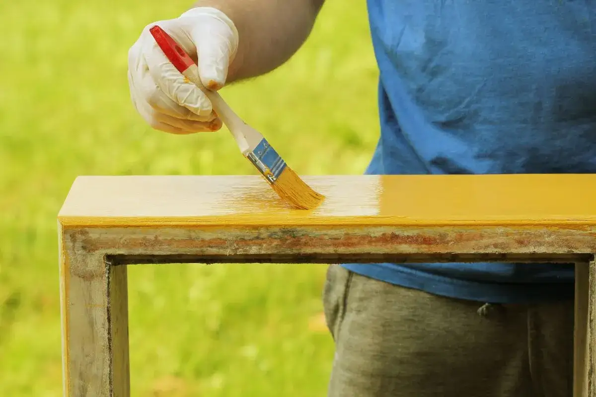 Persona con guantes blancos aplica pintura amarilla a un mueble.