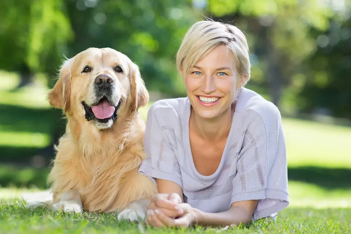 Uśmiechnięta kobieta i jej złoty retriever, jedne z najmądrzejszych psów, leżą na trawie w parku.