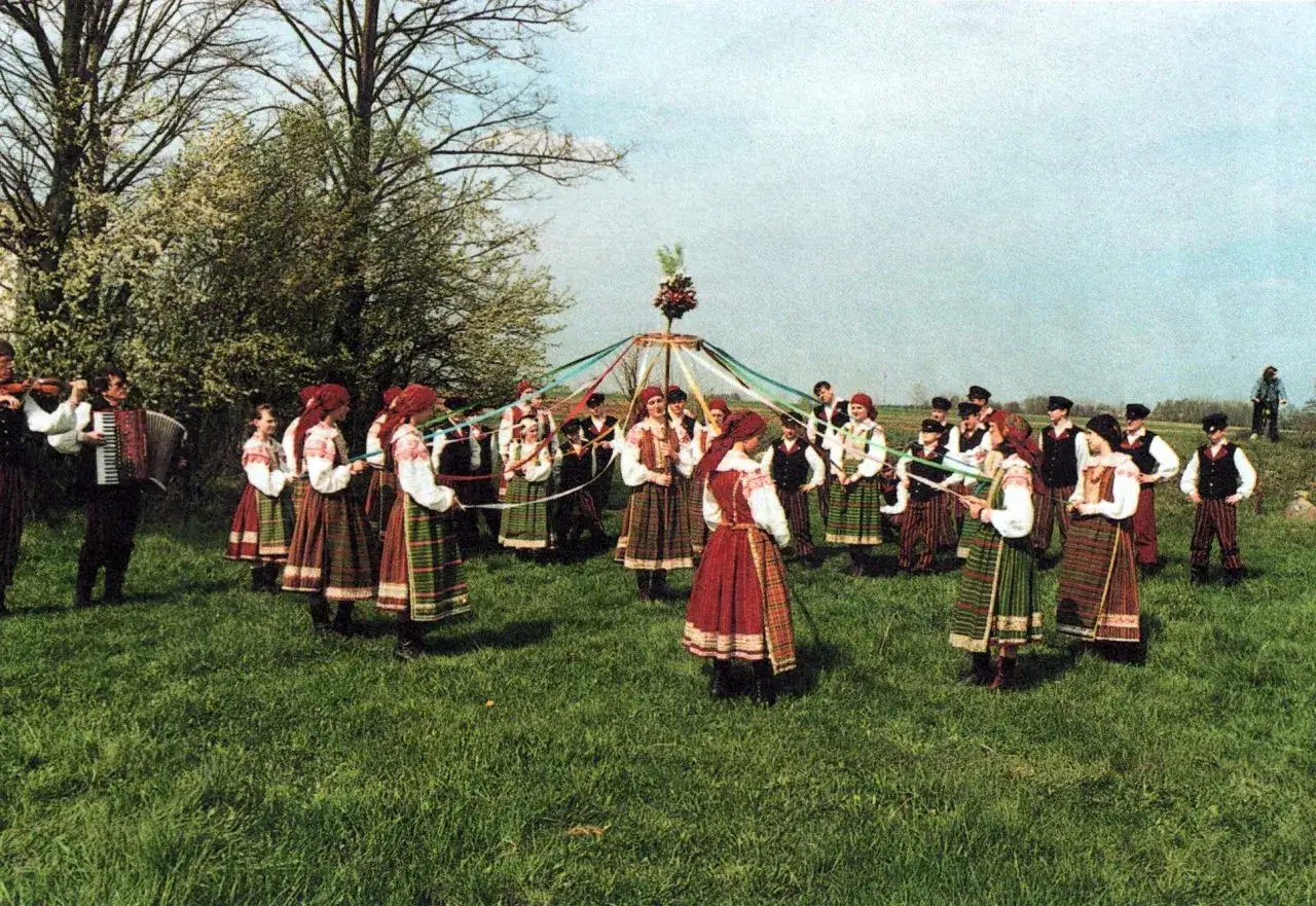 Ludzie w tradycyjnych strojach tańczą wokół udekorowanego słupa, celebrując zwyczaj ludowy nowe latko. Muzyka z akordeonu i skrzypiec towarzyszy radosnemu wydarzeniu.