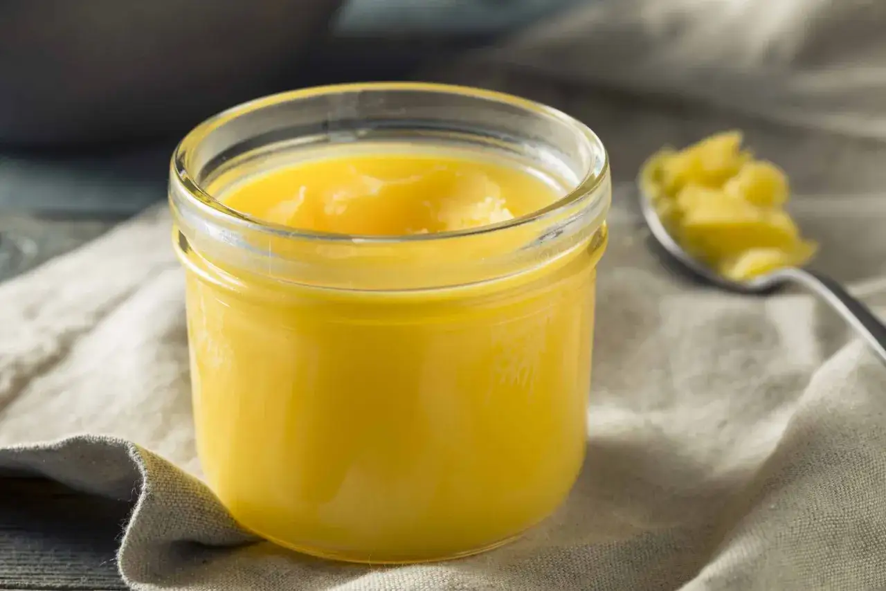 En burk med gyllene ghee, perfekt för ditt nästa ghee recept. En sked med ghee syns i bakgrunden.
