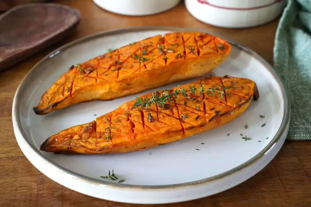 Deux moitiés de patate douce au four, grillées et parsemées de thym frais, sur une assiette blanche.