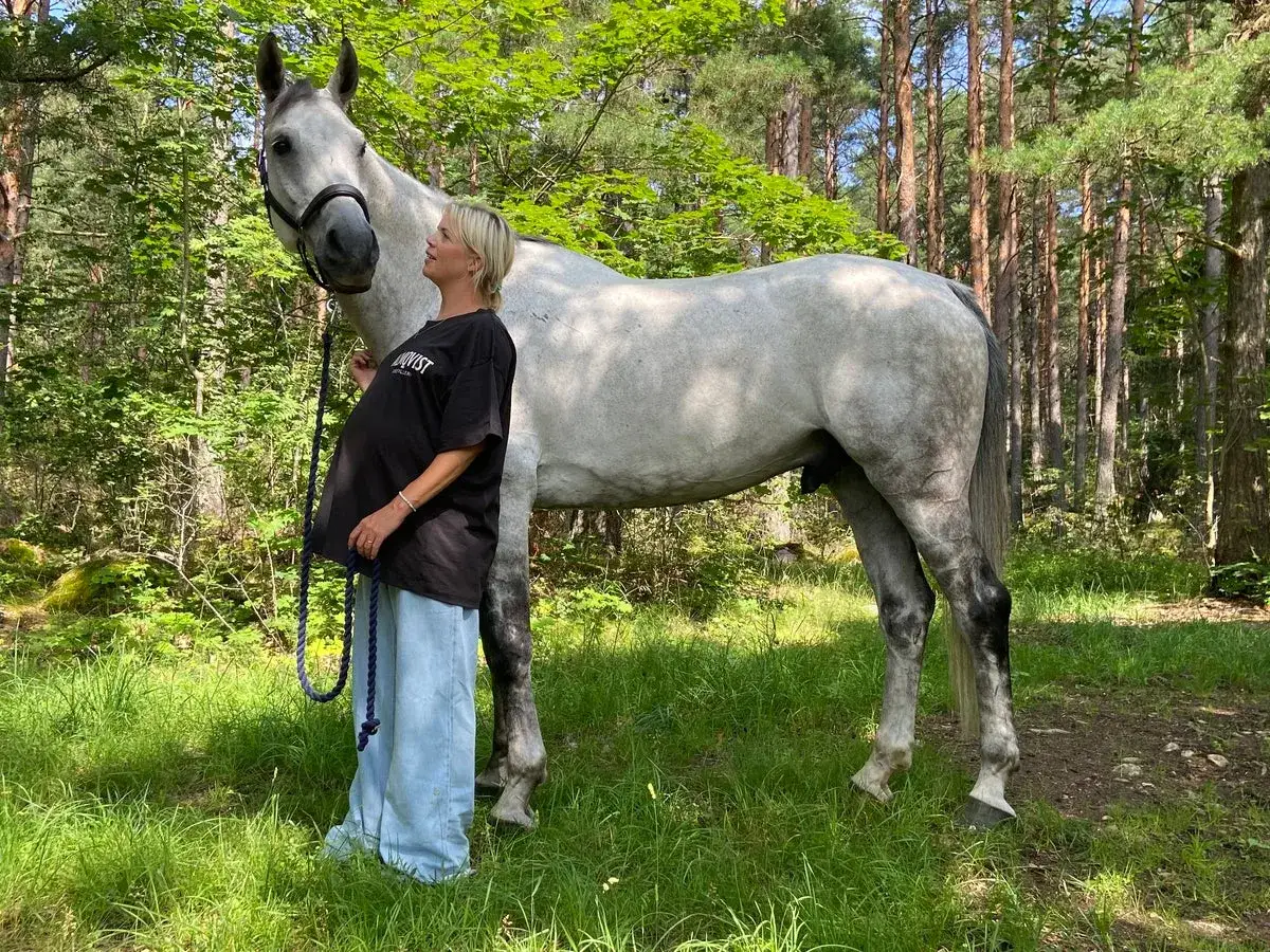 Gravid kvinna står bredvid en vit häst i skogen. Hon håller i hästens grimma, redo att rida.